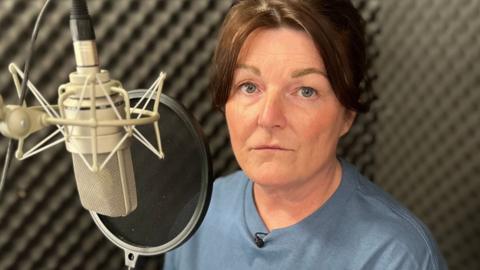 Gayanne sits in front of a microphone in a grey soundproofed recording booth, she has brown hair and wears a blue T-shirt