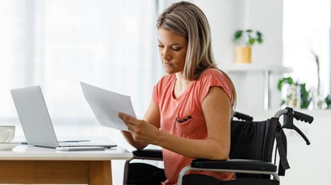 A woman sitting in a wheelchair in front of her laptop, looking at paperwork.