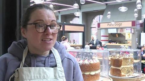 Linsay Rouston-Turner standing at her stall within a market hall. She is wearing glasses and a white apron and there are two large decorated cakes to the right of the shot.