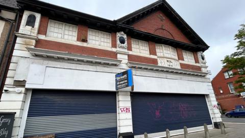 The outside of a boarded up shop which is a large brick building with a pointed roof. The ground level is covered by a white shop front and blue shutters. A "to let" sign is on the front of the of the building.