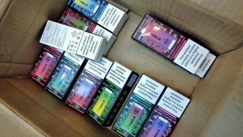 A cardboard box with 10 illegal vape boxes lying in the bottom. They are colourful designs with a picture of the rectangular vape on the front, along with a warning that the product is highly addictive.