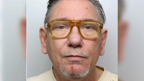 A police custody photo of a man with grey hair and grey stubble, wearing a pair of yellow glasses and a pale T-shirt. He's looking straight at the camera and has a plain white wall behind him.