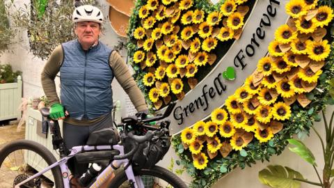Stephen Aslin, wearing a white helmet and blue body-warmer, poses with his bike in front of a floral Grenfell memorial