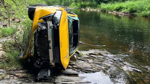 The yellow car is lying on its side next to the River Dart. The windscreen is cut away and smashed, the bumper is ripped away and its wheels are askew. 