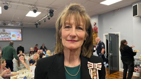 Anna Turner, a woman with dark blonde shoulder length hair and a fringe is smiling at the camera and wearing a black blazer with green top underneath and a gold chain. on her shoulder is a white and red bow