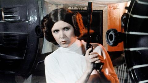 Actress Carrie Fisher, wearing a white costume and two hair buns, holds up a black metal gun on a sci-fi filmset.