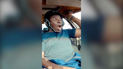 Clive Myrie looks elated in a car wearing a helmet and holding on to a handle