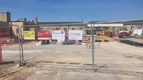 Chippenham Rail Station, with a building sire in front - large metal gates with piles of materials and machinery behind. Sunnny day.