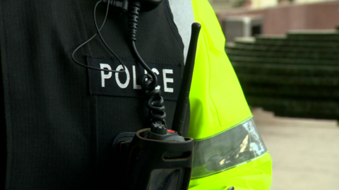 A closeup of a police officer's uniform. The hi-visibility sleeve can be seen, as can a radio in a pocket. Police is written just above the pocket.