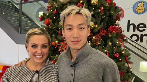 Amy Dowden and Carlos Gu in the foyer of BBC Birmingham with a staircase and a Christmas tree behind them. Dowden is wearing a top with a black and white check pattern and Gu is wearing a grey jumper. They are both smiling at the camera, looking happy.