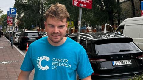 A man with brown hair and a beard with a blue tshirt which reads cancer research uk in a street with a black car behind him