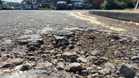 A close up photograph of a pothole. There's lots of loose stones and patchy tarmac. There's two double yellows in the background next to the pavement. Out of focus at the top of the photograph is cars and houses from the street.