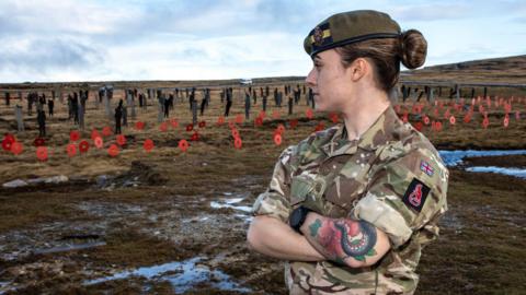Rhian Brown looks at a display of poppies. Rhian is wearing military fatigues and a beret and has her arms crossed looking away from the camera.