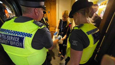 Two police officers have their back to the camera while taking a swab of  a sample. A lady with blonde hair and a black outfit is stood opposite holding the sample.