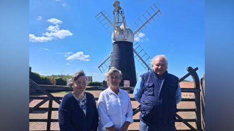 Two women and a man standing in front of a windmill. The windmill is black and has four main sails and a fantail to the rear.
