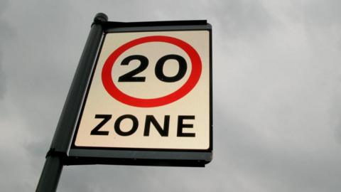 A 20mph zone sign on a pole with a grey sky as a back drop.