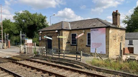 A small stone brick building sits behind railway lines on a sunny day. There's a railway crossing to the right with trees. 