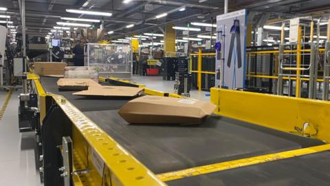 Amazon parcels roll along a black conveyer belt with yellow barriers inside a warehouse building 