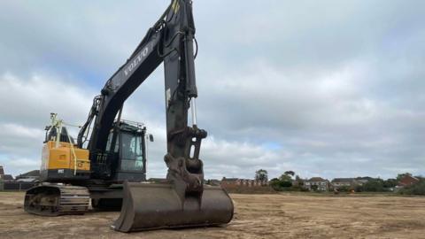 A large Volvo digger, with a glass cab, yellow body and black crane arm, stands on bare earth, with it's bucket on the ground ready to dig.