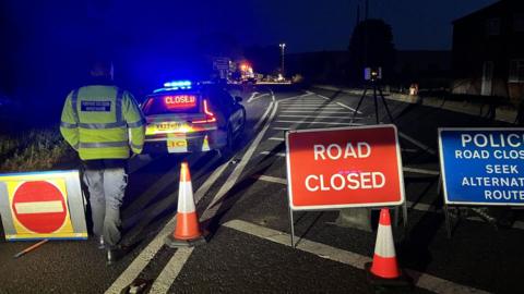 A main road has been cordoned off by police in the evening. There are road closed signs, with traffic cones and a police car blocking off the road and a forensic collision investigator.