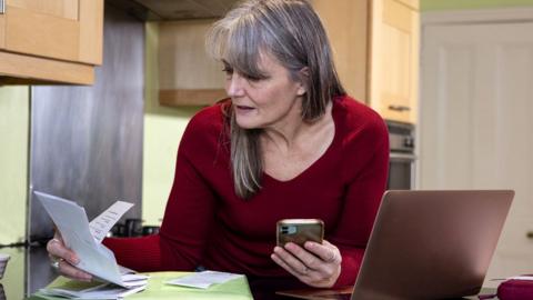 A middle-aged woman wearing a red top stands in her kitchen next to a laptop on a table whilst holding her mobile phone and looking at bills in a green folder.