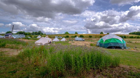 A number of tents are in the middle of the photo, including the largest one, which is white and green. A van is also in the centre and the sky, including grey clouds, features in about half of the image.