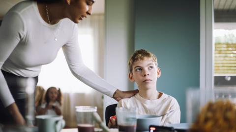 A boy looking away from a woman with her hand on his shoulder. He is sitting at a kitchen table.