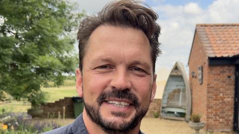 Jimmy Doherty smiles at the camera. He has short brown hair and a brown beard. He wears a blue shirt.