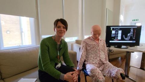 Occupational therapist Ruth Alecock sitting in a room next to robot Mona. There is a screen behind them which shows impact of touch on the robot. 