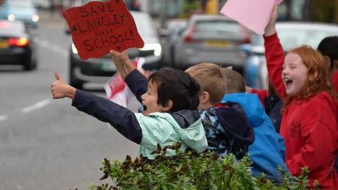 A group of young boys facing cars on the road with placards. One boy is holding up a handmade sign which reads Save Langley First School in one hand and holding his thumb up with his other.