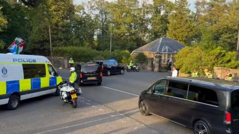 A police van and motorbike parked in front of black vans driving through a country estate gates.