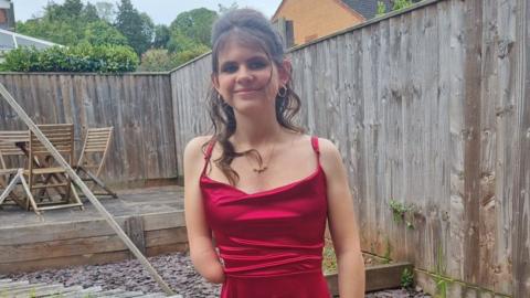 Paige Cox is standing in a back garden wearing a red dress. Her hair is done up. She is smiling.