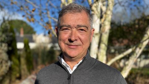 Rob Jones, a man with short grey hair and stubble. He is wearing a dark grey quarter neck jumper over a white shirt. He is looking directly ahead. He is outdoors. Behind him are tree trunks, greenery and a blue sky.
