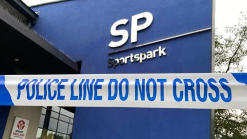 Police tape in front of the Sportspark building. The part of the building in vision is a large blue wall with the silver lettering "SP" and "Sportspark".