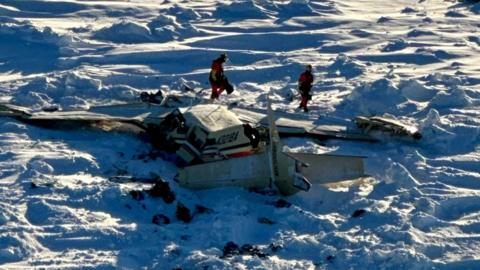 The wreckage of the plane, as seen from the air, surrounded by snow and debris as two people walk around it