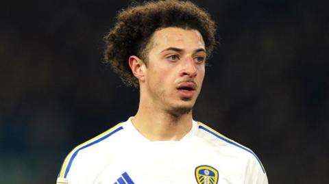 A close-up of Ethan Ampadu on a football pitch. It is dark behind him and he is wearing a white Leeds United football strip with yellow and blue stripes on the shoulders. He is looking to the right of the camera and his mouth is slightly open with sweat on his forehead.