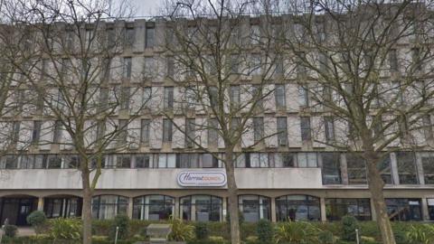 A tall concrete building with large windows, partly hidden behind bare trees. A sign above the entrance reads "Harrow Council, London.