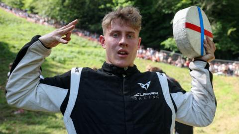 Tom Kopke holding a wheel of cheese. He has short blonde hair and is wearing a black and white jacket with a Cupra racing badge. He is stood at the bottom of Cooper's Hill after winning a cheese rolling race.