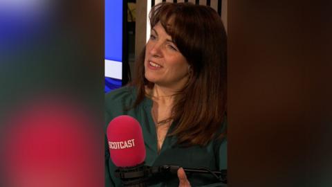 Catriona MacPhee in a green shirt speaking into a pink microphone with the word 'Scotcast' on it.