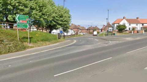 A road junction with green road signs and white road markings. There are trees to the left of the image and houses in the background to the centre and right.