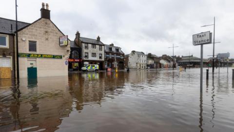 The Whitesands area of Dumfries in deep brown water from the nearby River Nith. A taxi stance sign is partially submerged and there are boarded-up shops and a police van in the distance.