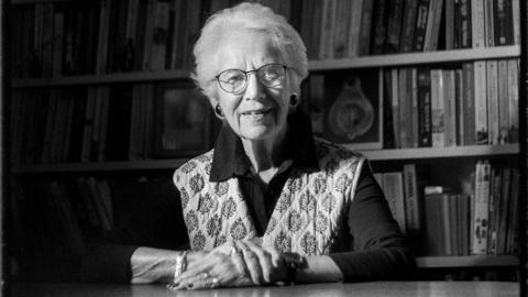 A black and white portrait of an Eva Clarke with short white hair and glasses, sitting at a table with her hands clasped, in front of a bookshelf filled with books.