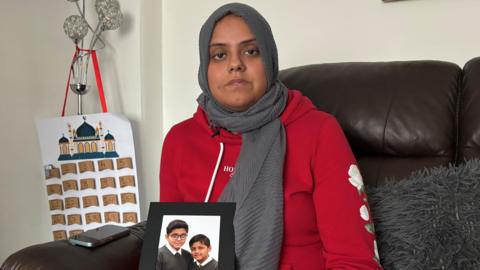 A mother wearing a grey headscarf and red jumper is holding a photograph of two children. She is sitting on a brown leather chair with a grey furry cushion next to her.