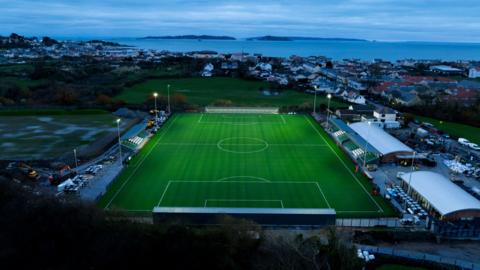 A night time aerial view of the football stadium, with the floodlights on and houses and the sea in the distance.