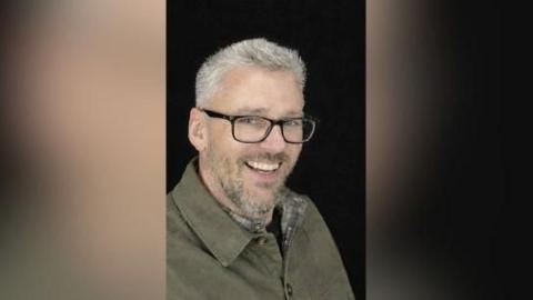 Craig Hurcombe smiles at the camera. He wears a green collared jacket and black rimmed glasses. He has short grey hair and a beard. 