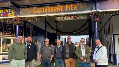 Seven men standing in front of Eastbourne Pier.
