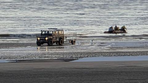A 4x4 vehicle waits at the shore for an inflatable RNLI lifeboat with crew members in it in the sea.