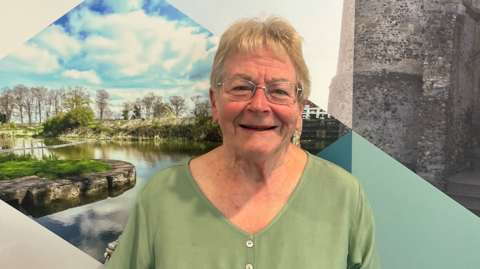 Sue Dillon, who has a green, buttoned-up top and short, white hair, is also wearing silver-framed glasses 