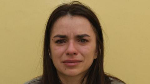 Police photo of Vanya Gaberova who is pictured with black hair which falls slightly below her shoulders. She is in front of a dark yellow background and appears to be crying. 