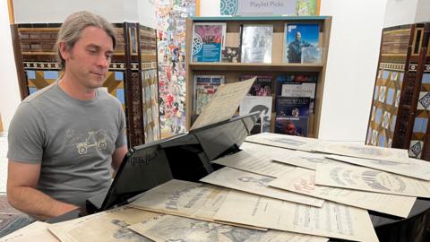 A man with long tied-back brown and grey hair sits at a piano which is covered in sheet music. He is wearing a blue T-shirt.
